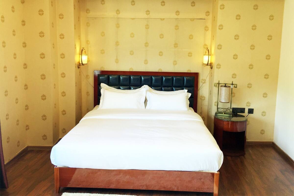 A hotel room with a bed, wooden headboard, patterned wallpaper, and two wall-mounted lamps.