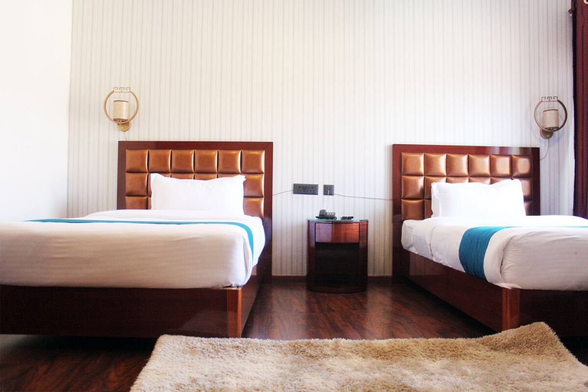 A hotel room with two beds, a wooden headboard, a small bedside table, and a wall-mounted lamp.