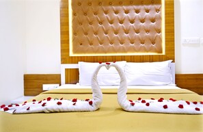 A hotel room with a tufted headboard, white bedding, and two swans made of towels with rose petals. - Hotel TR Residency (Puducherry)