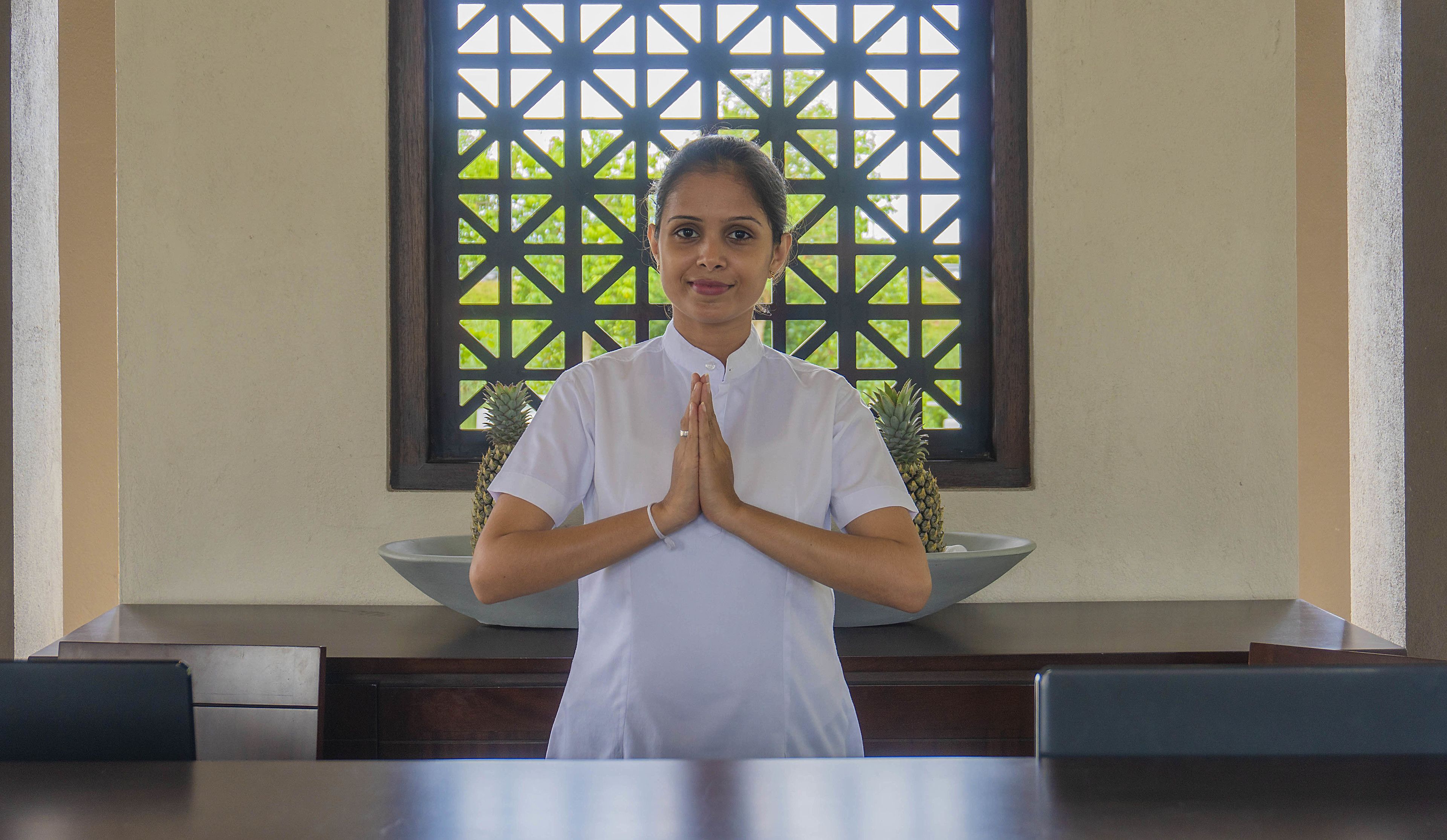 A person in a white shirt standing in a room with a geometric patterned window, a bowl, and a table.