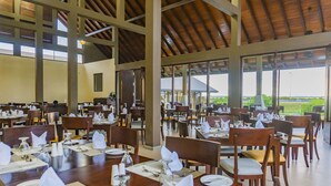 Un comedor amplio con varias mesas dispuestas para recibir a los huéspedes, grandes ventanas que ofrecen vistas al exterior y un techo alto con vigas de madera.