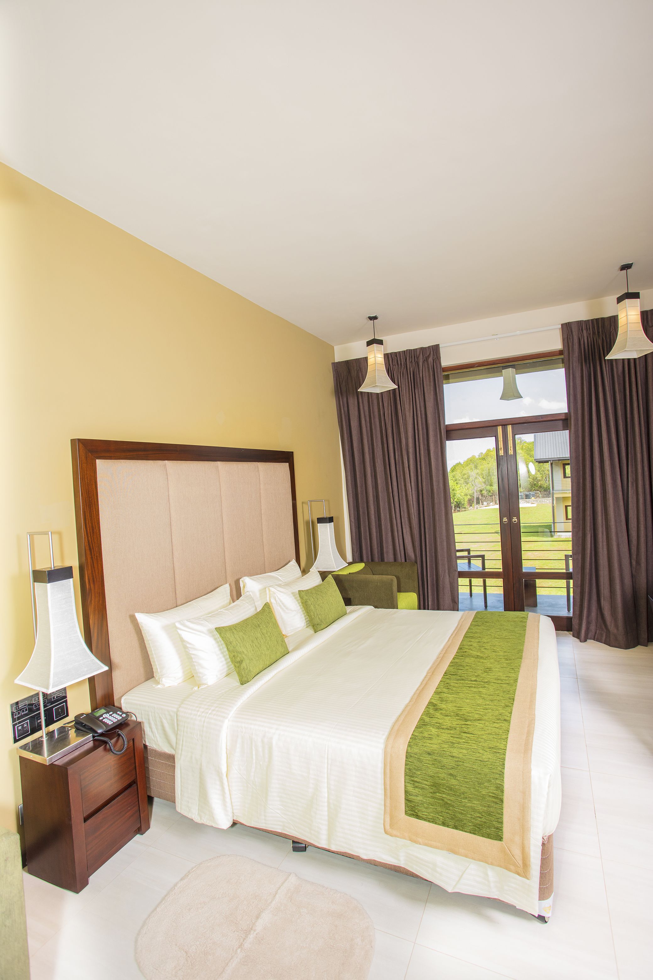 Habitación de hotel con una cama grande, una mesita de noche con lámpara, un teléfono y vistas al exterior a través de un ventanal.