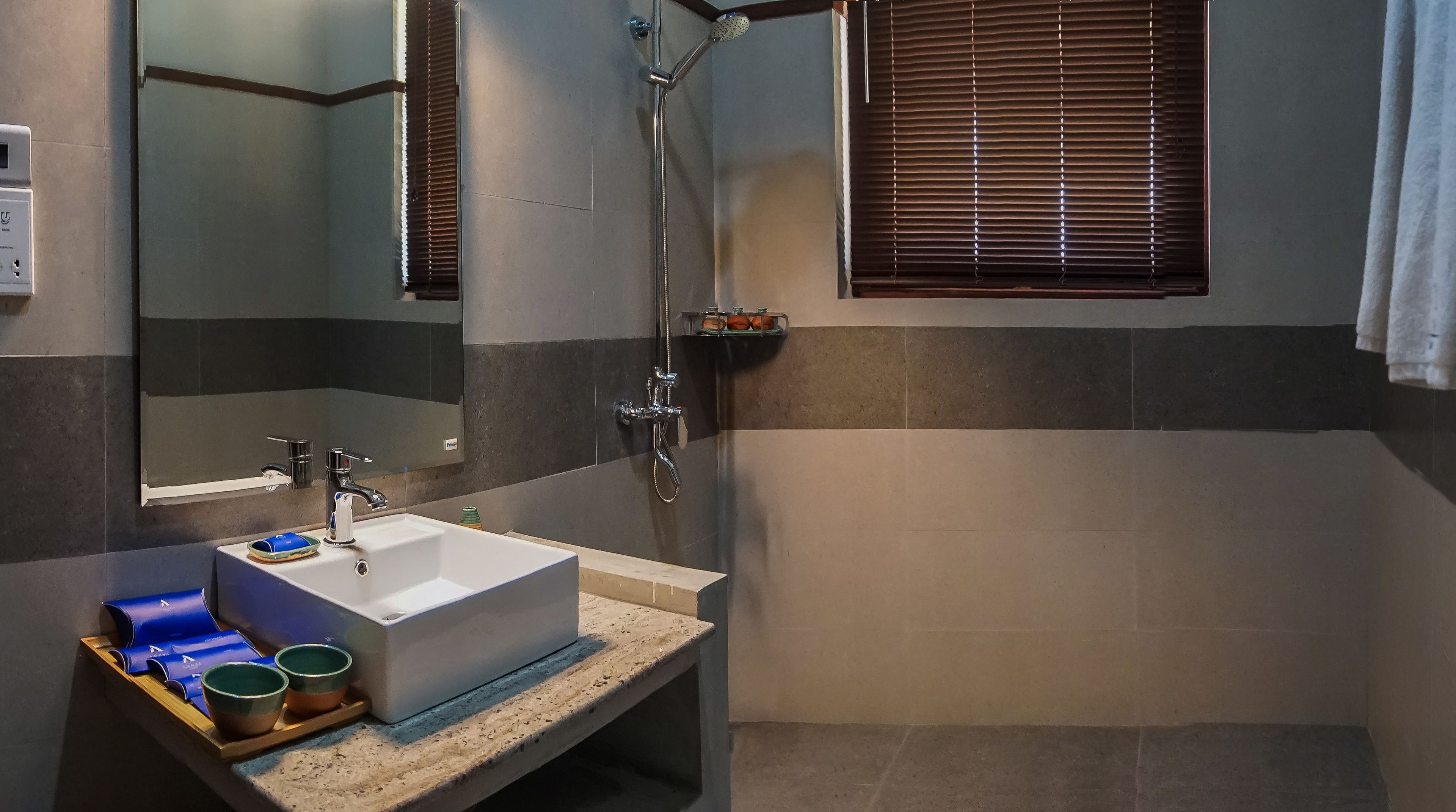 Een badkamer met een wit wastafelmeubel, een spiegel, een douche en een raam met jaloezieën.