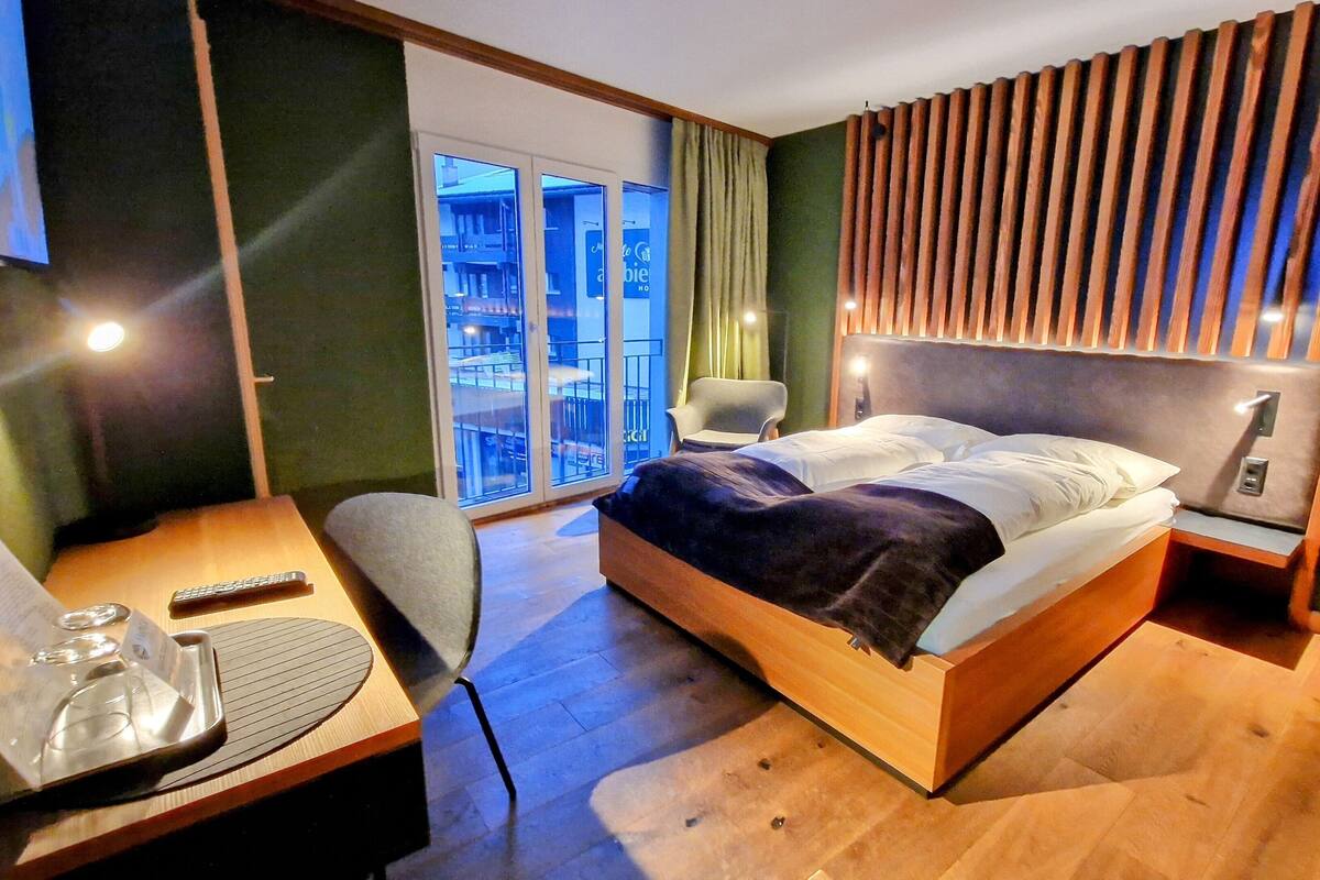 A hotel room with a bed, a desk with a chair, a lamp, and a view of the outside balcony.