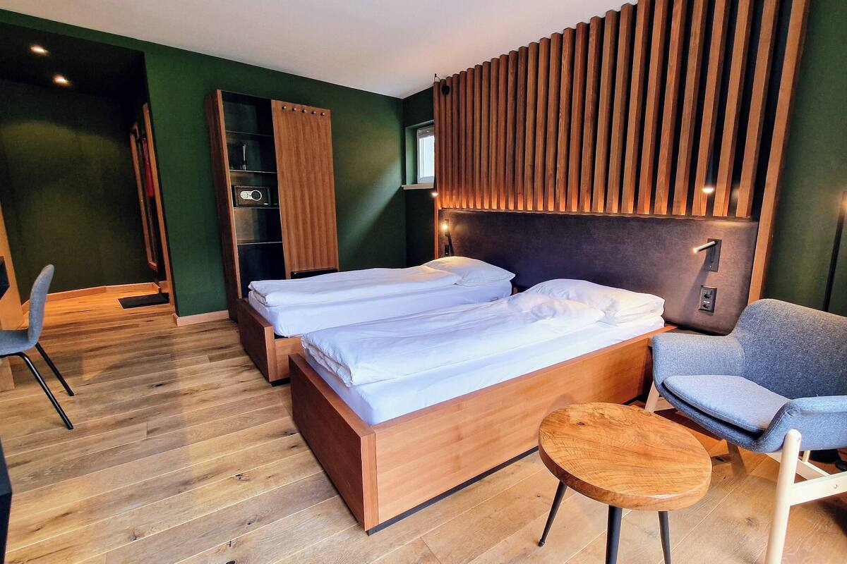 A modern hotel room with two single beds, a wooden headboard, a grey armchair, a small wooden table, and a green wall.