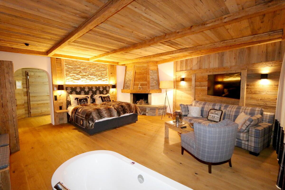 A wooden-floored room with a large bed, a sofa, a chair, and a fireplace.