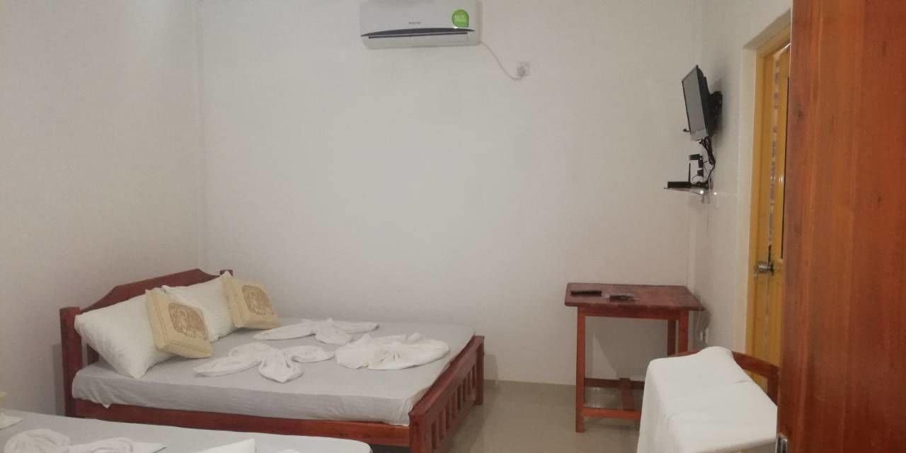 A small, clean room with a single bed, a bedside table, a TV mounted on the wall, and a wooden door.