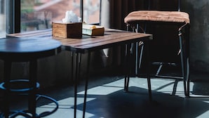 A room with a large window, a wooden table, a cushioned chair, and a small wooden box. - Samantan Hotel at Nimman (Chiang Mai)