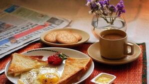 Une table mise pour le petit-déjeuner avec des toasts, des œufs, des fraises et une tasse de café, ainsi qu’un journal et un vase de fleurs.