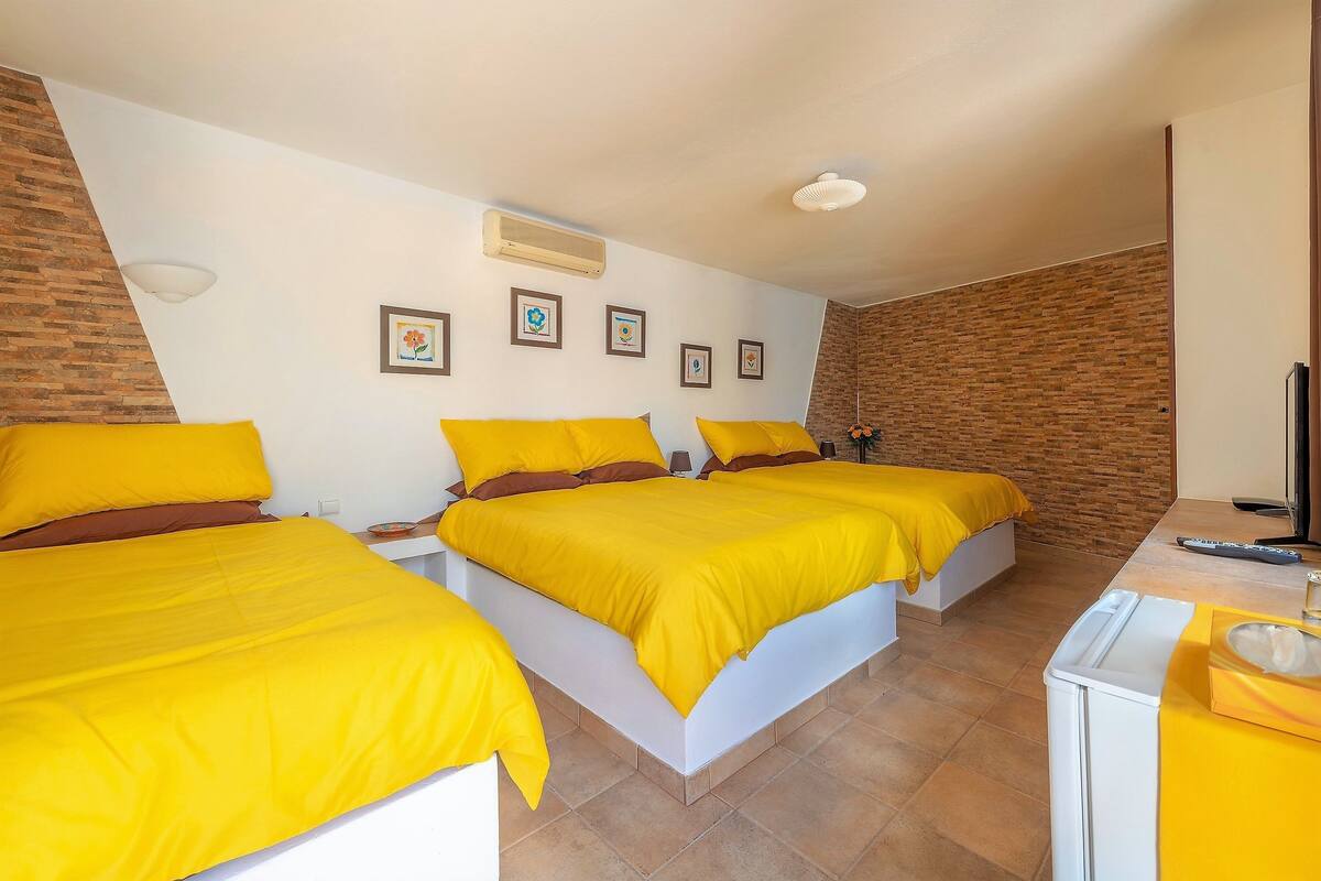 A hotel room with two beds, each with a yellow bedspread and brown pillows. There is a small table with a yellow tablecloth and a white appliance on it. The room has a brick wall and a white ceiling with a light fixture.