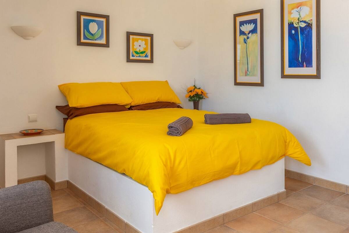A bed with a bright yellow bedspread, a small bedside table with a bowl, and three framed artworks on the wall.