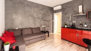 A modern living room with a grey sofa, red cushions, a small side table, and a red kitchen area with cabinets and a countertop. - Guest house Roma (Rome)