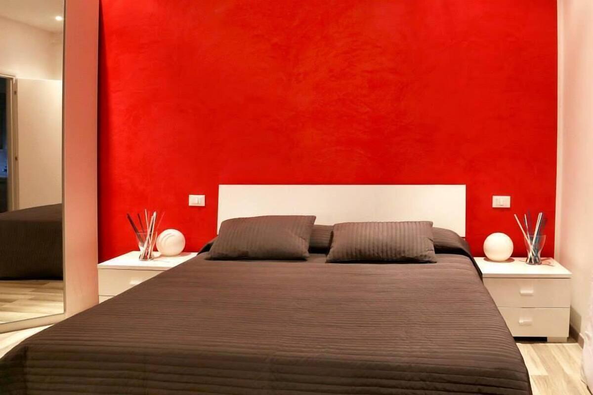 A modern bedroom with a large bed, a red accent wall, and bedside tables with decorative items.