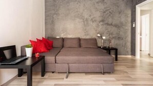 A modern bedroom with a grey sofa bed, a small table with a laptop, a potted plant, and a nightstand with a lamp. - Guest house Roma (Rome)