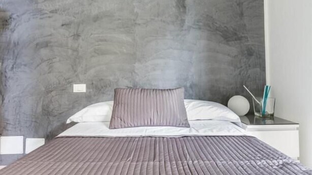 A bedroom with a grey textured wall, a bed with a grey and white duvet, and a bedside table with a glass containing a straw. - Guest house Roma (Rome)