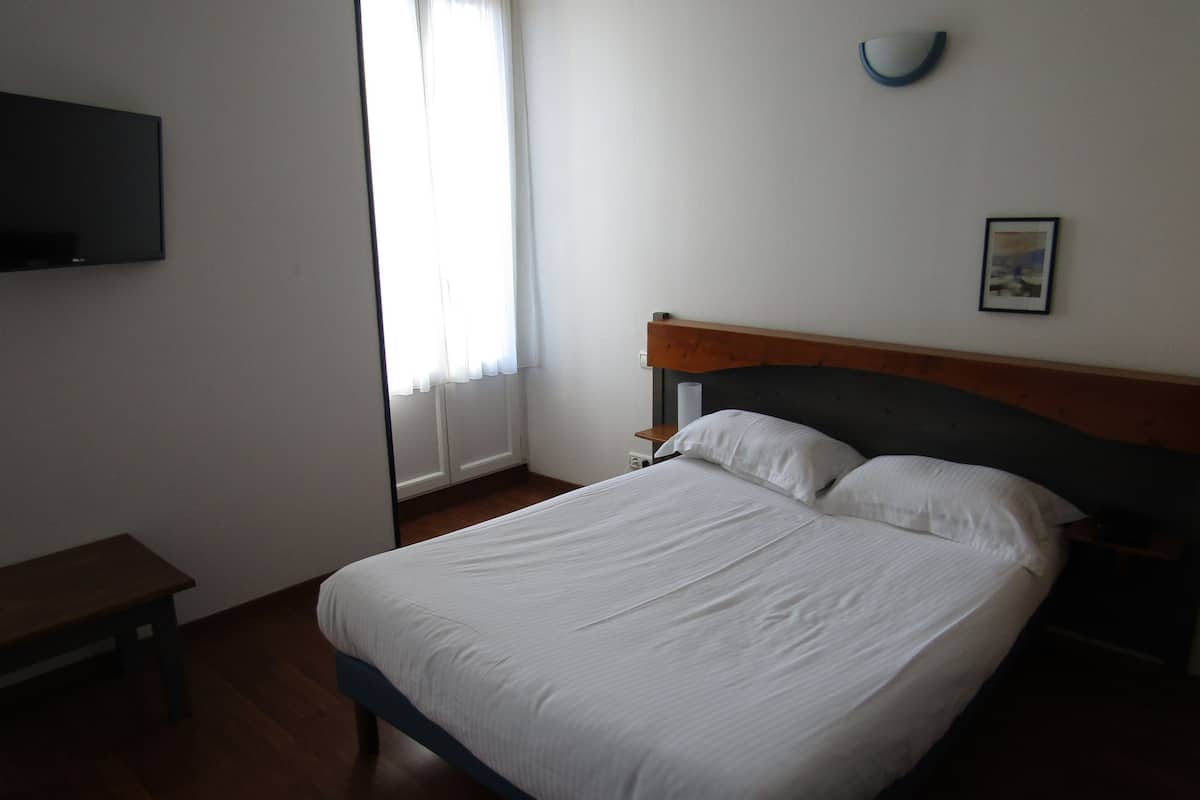 Ein ordentlich bezogenes Bett mit weißen Leinentüchern in einem Zimmer mit Holzboden, einem Nachttisch, einem an der Wand befestigten Fernseher und einem Fenster mit Vorhängen.