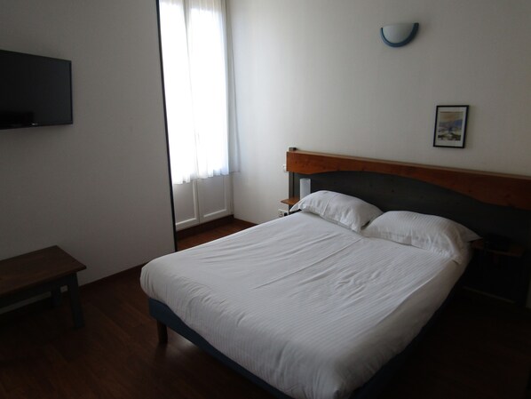 A neatly made bed with white linens in a room with wooden flooring, a bedside table, a wall-mounted TV, and a window with curtains.