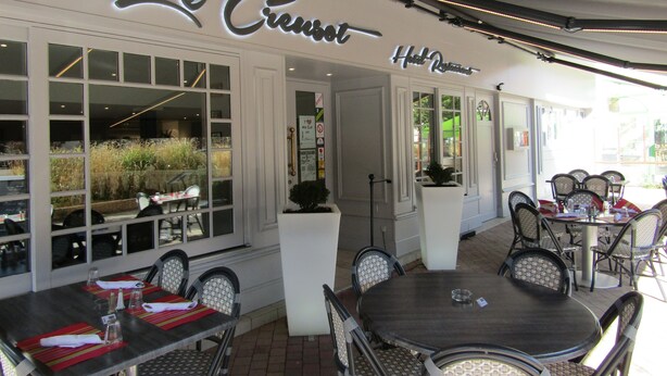 An outdoor seating area with tables and chairs, a sign reading "Le Creusei Hotel Restaurant," and a view of the street through large windows. - Le Creusot Hôtel (Le Creusot)