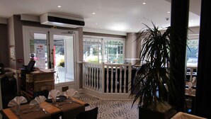 A dining area with a table set for two, a potted plant, and a view of the street through a glass door. - Le Creusot Hôtel (Le Creusot)