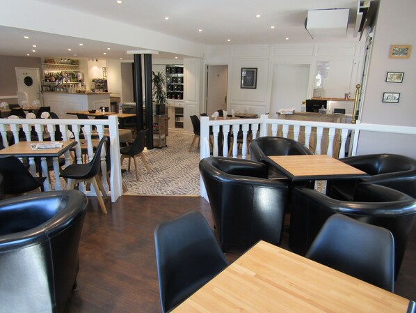 Un restaurante con mesas de madera y sillas negras, una zona de bar con botellas, y una barandilla blanca que separa el comedor de la barra.