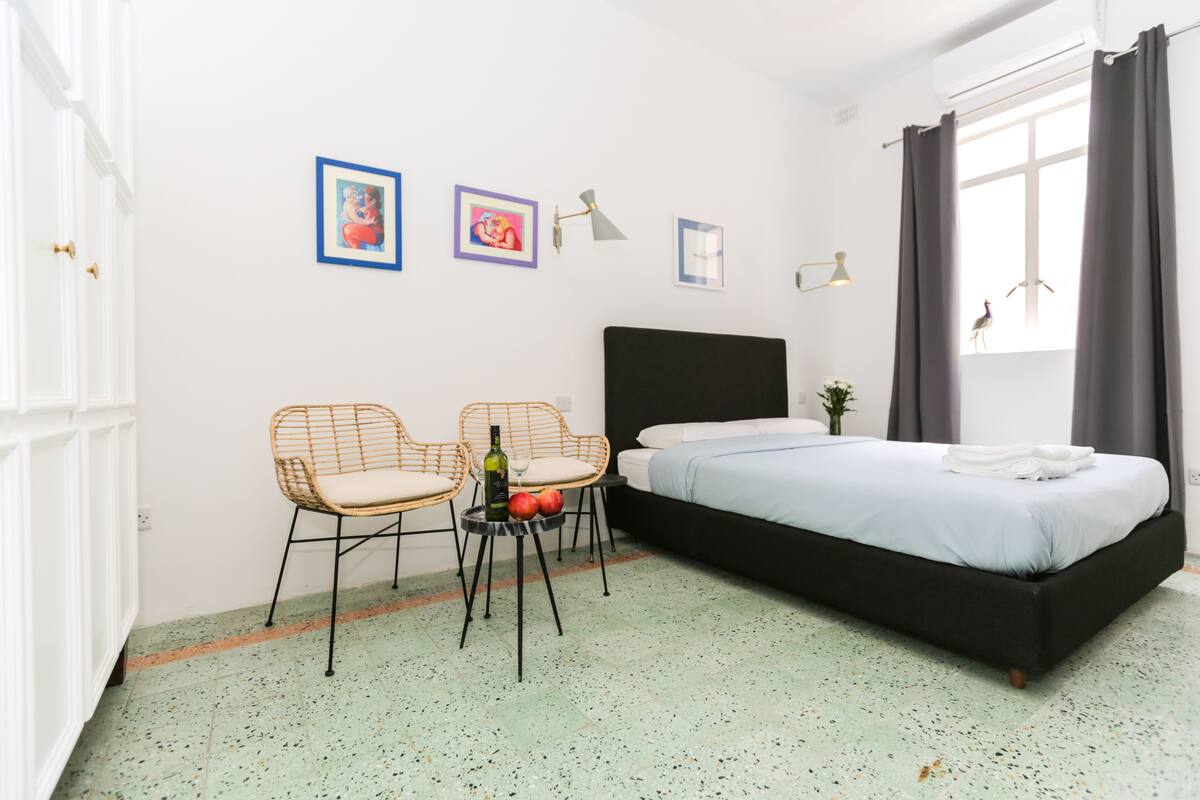 Un dormitorio con una cama, dos sillones de mimbre, una mesita con una botella y manzanas, y una ventana con cortinas.