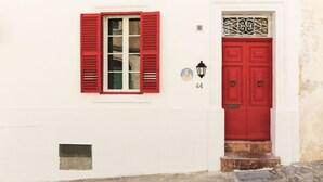 A white building with a red door and window shutters, a small window with a metal grate, and a number '44' on the wall. - House of Pomegranates (Sliema)