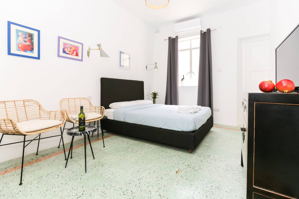 Un dormitorio con una cama, dos sillones de mimbre, una mesita con una botella y un aparador con manzanas.