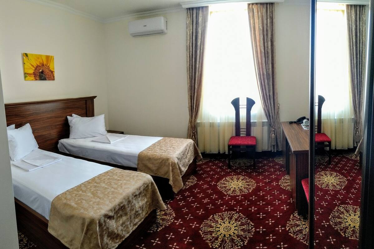 A hotel room with two single beds, a wooden headboard, a red patterned carpet, and a small table with a chair.