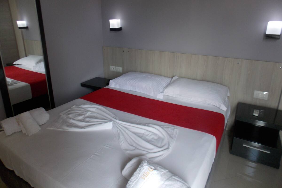 Habitación de hotel con cama, mesita de noche, espejo y lámpara de pared.