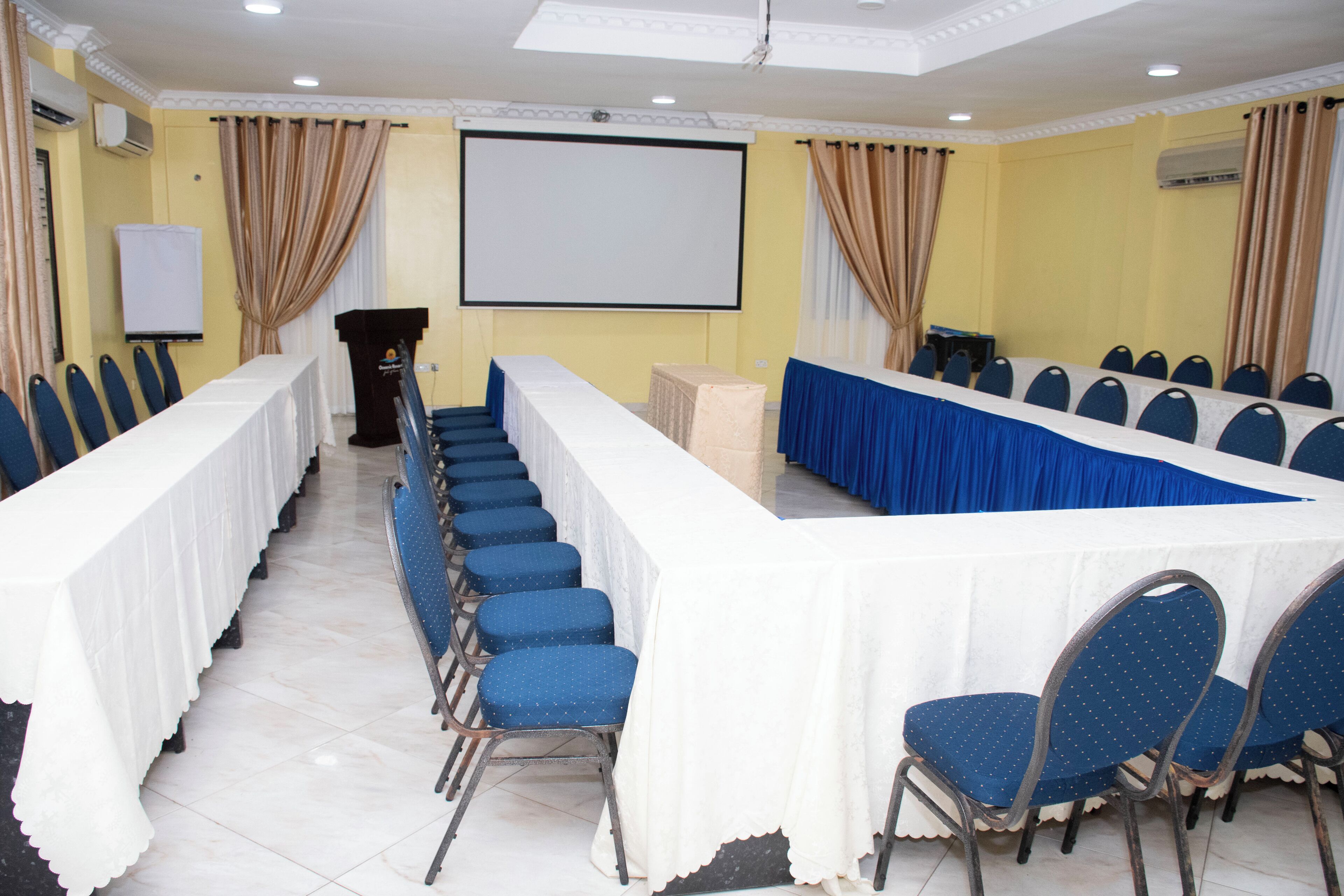 Un salón de conferencias con una larga mesa, sillas azules, un atril y una pantalla de proyección.