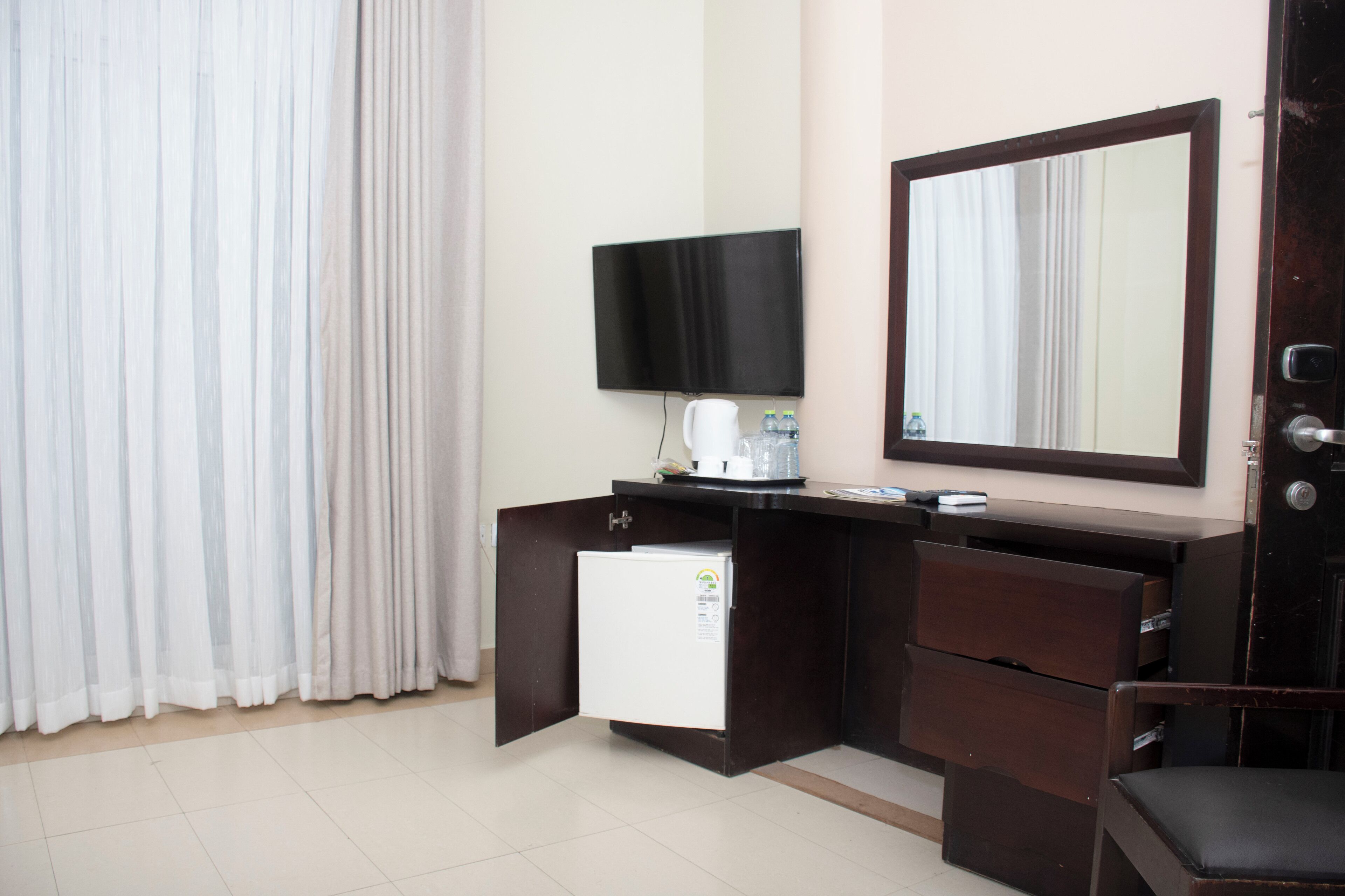 Quarto de hotel com escrivaninha de madeira escura, mini-frigorífico branco, televisão de tela plana, espelho e uma cadeira.