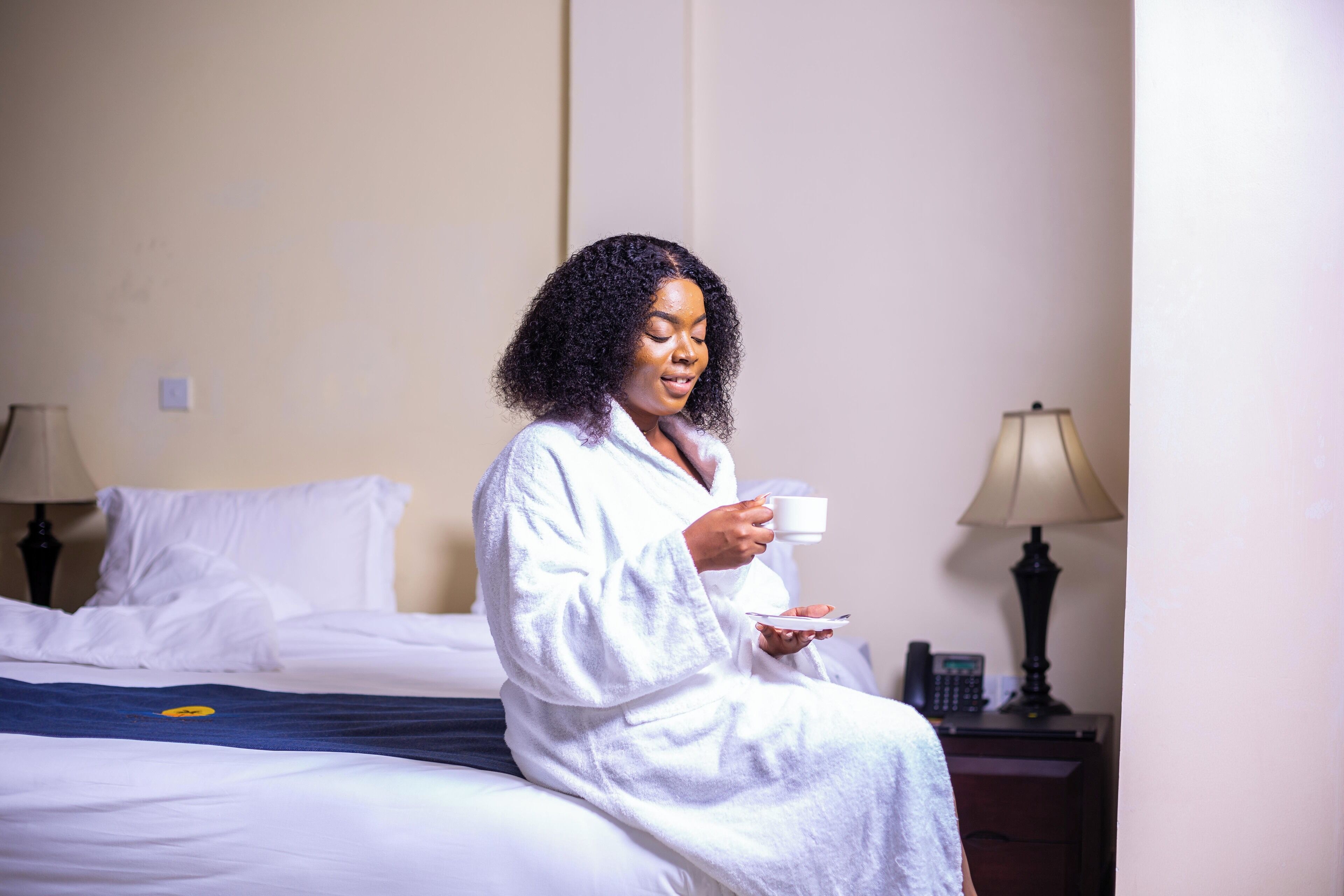 A person sitting on a bed in a hotel room, holding a cup and a plate.