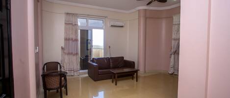 A living room with a brown leather sofa, a wooden coffee table, and a chair. There is a ceiling fan and an air conditioning unit on the wall.