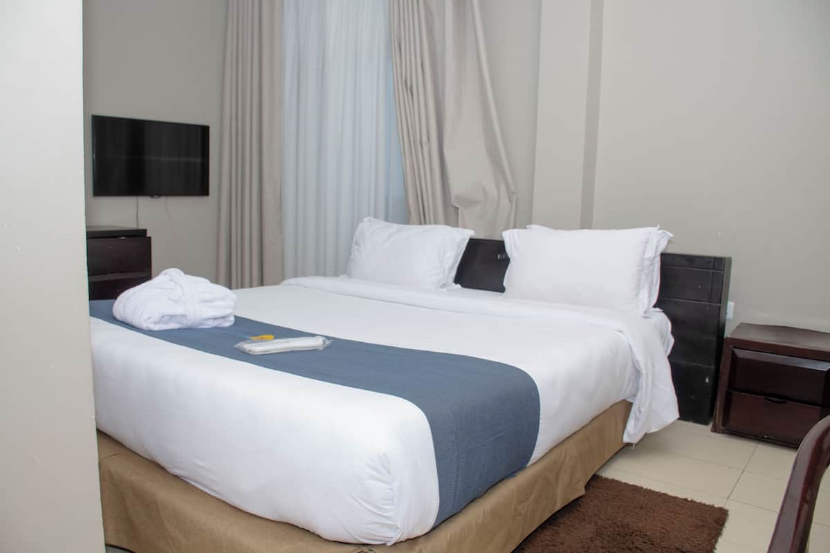 A neatly made bed with white and blue bedding, a flat-screen TV on the wall, and a brown rug on the floor.