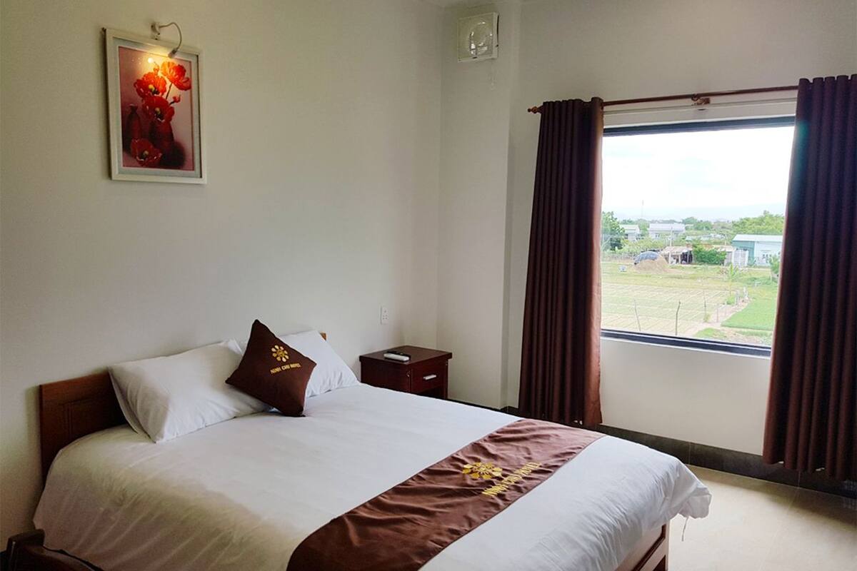 A hotel room with a bed, a nightstand, a framed picture on the wall, and a window with curtains.