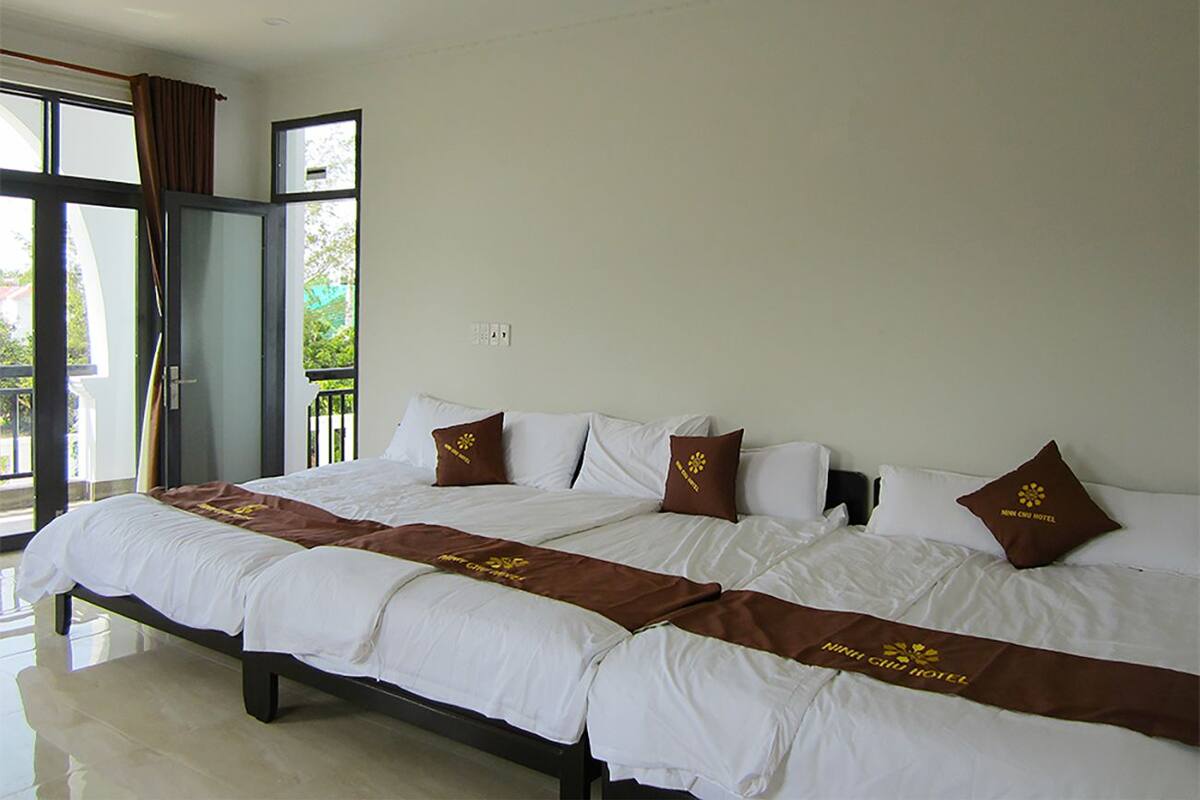 A double bed with white linens and brown decorative pillows, a large window with curtains, and a view of greenery outside.