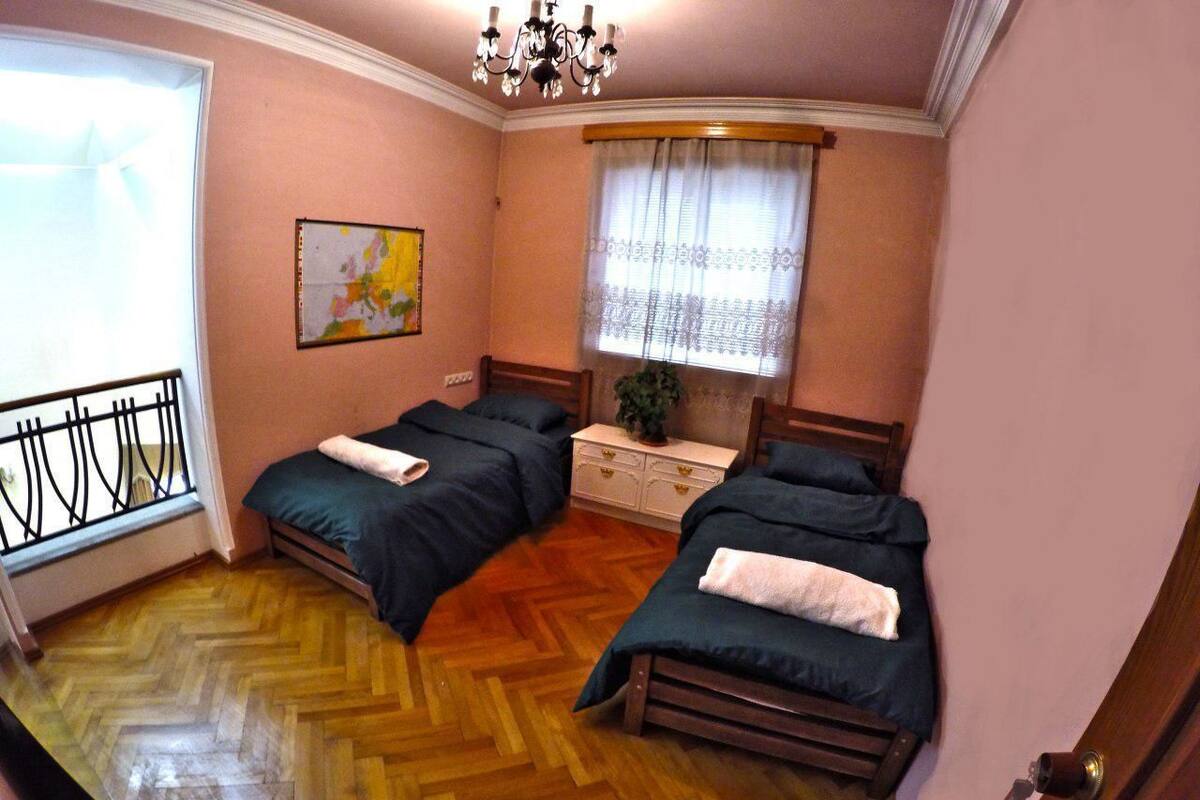 A room with two beds, a wooden herringbone floor, a window with a curtain, and a chandelier.