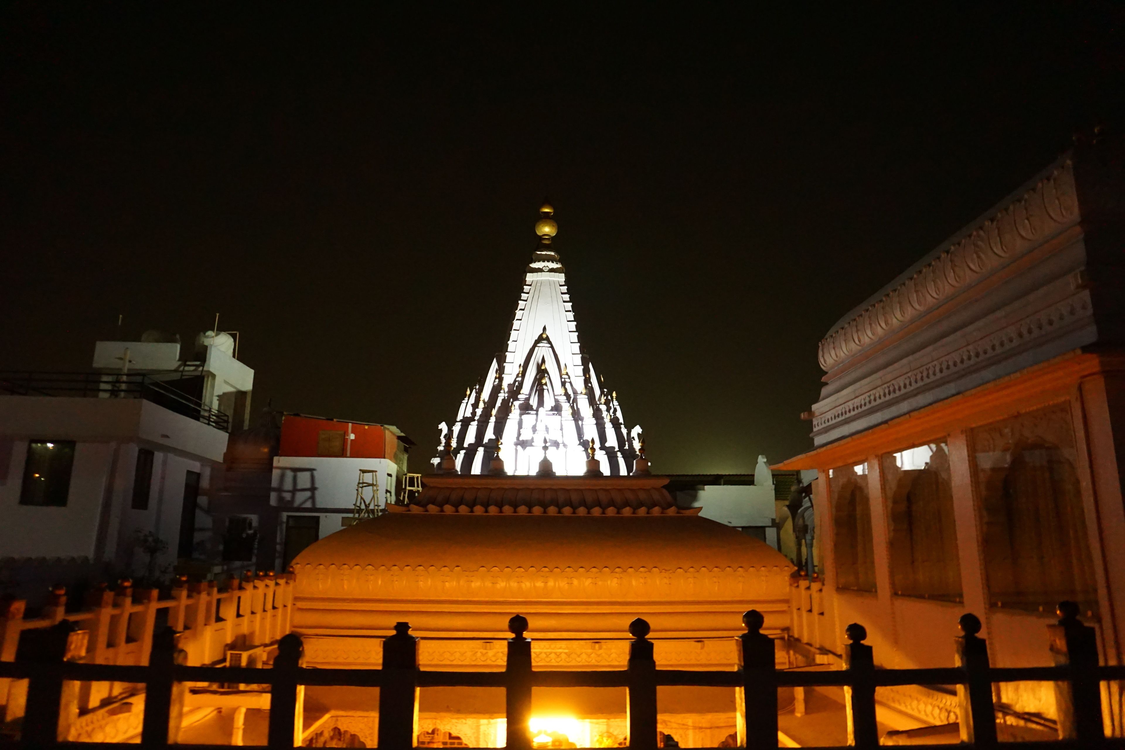 A night view of a temple with a prominent spire and a large central dome, surrounded by a fence and adjacent buildings.