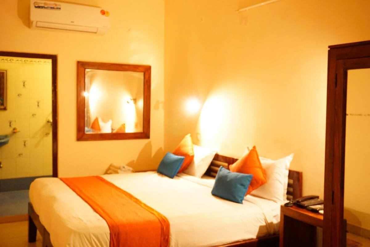 A hotel room with a bed, pillows, a bedside table, a mirror, and an air conditioning unit.