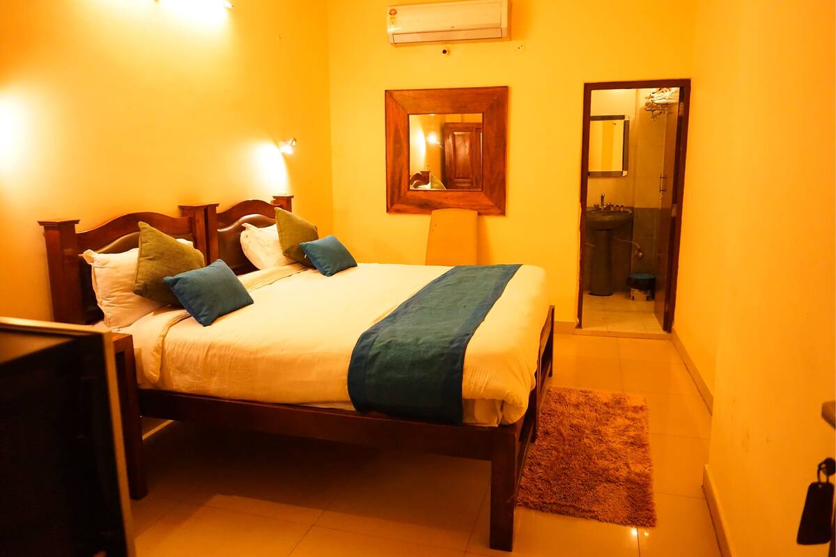 A neatly made bed with pillows and a blanket, a wooden headboard, a bedside table, a mirror, and a bathroom visible through an open door.