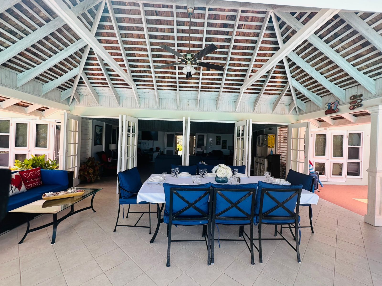 A covered patio with a dining table set for six, surrounded by blue chairs, and a ceiling fan above.