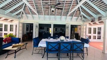 A covered patio with a dining table set for six, surrounded by blue chairs, and a ceiling fan above.