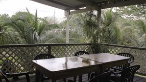 Un patio al aire libre con mesa y sillas de madera, rodeado por un cerramiento de celosía, con vistas a un exuberante jardín con palmeras.