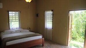 Un dormitorio con cama de madera, dos ventanas con cortinas blancas y vistas a un jardín por una puerta abierta.