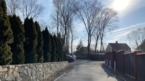 Une allée étroite, bordée d’un côté par un mur de pierres et de l’autre par une clôture en bois, mène à un quartier résidentiel où se trouvent des maisons et des voitures garées.