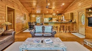 A log cabin interior with a living room featuring a leather sofa, a coffee table with newspapers, and a dining area with a table and chairs. - 3 Bedrooms, 1 Flex Room, 3 Bathrooms!  Hot Tub, Wifi & Pet Friendly!  Great Getaway For Any Season! (Huddleston)