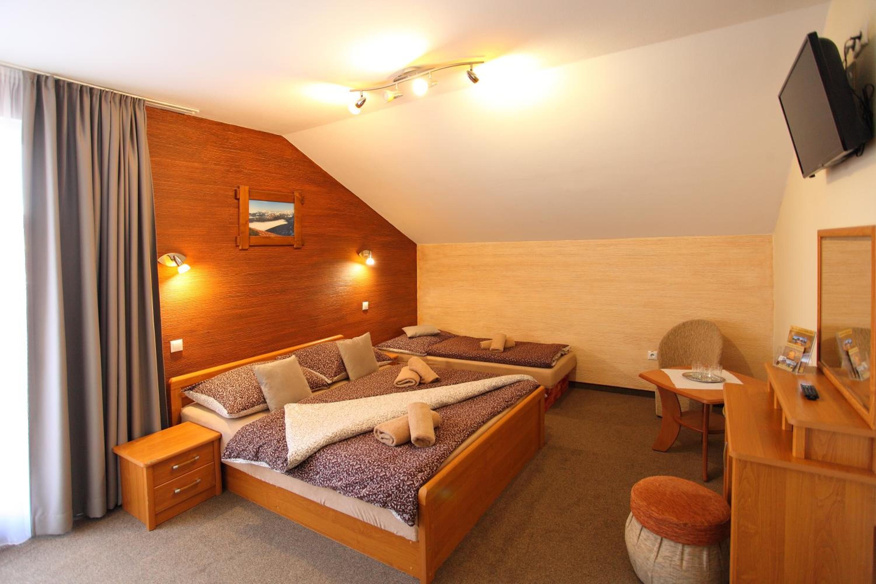 A bedroom with a wooden headboard, a bed with a patterned bedspread, a bedside table, a desk with a chair, and a flat-screen TV mounted on the wall.