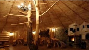 Un interior rústico de madera con techo alto, chimenea y un gran tronco de árbol como elemento central.