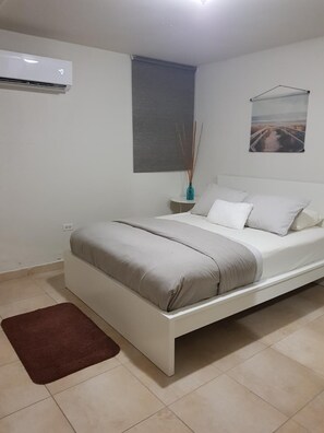 A bedroom with a bed, a nightstand, a wall-mounted air conditioner, a window with a blind, and a framed picture on the wall. - COMFORT /FANTASTIC,NEW COZY APART, 2 BEDROOM,NEAR AIRPORT (CAROLINA)