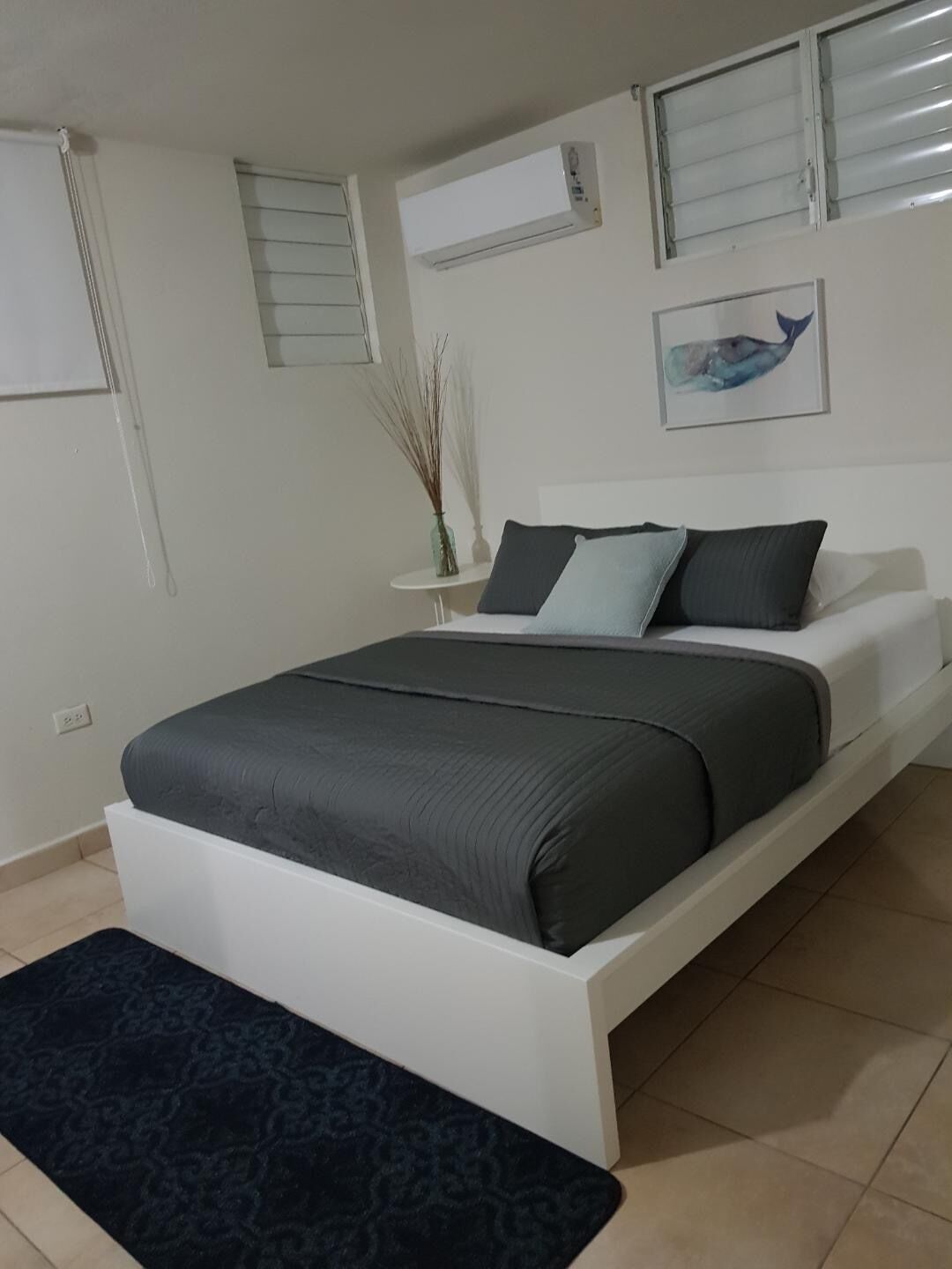 A bedroom with a bed, a nightstand, a wall-mounted air conditioner, and a framed picture.